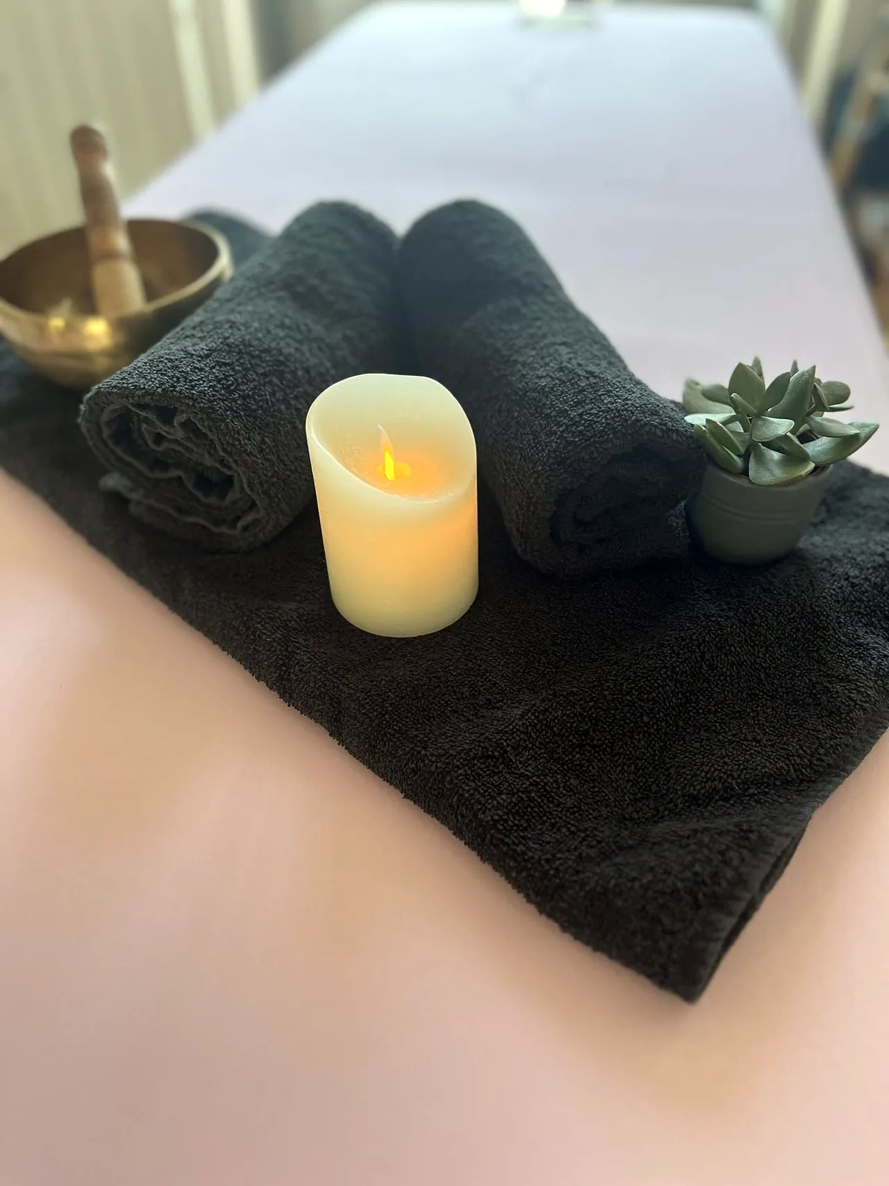 Des serviettes noires roulées, une bougie et un bol chantant en laiton, posés sur un drap rose